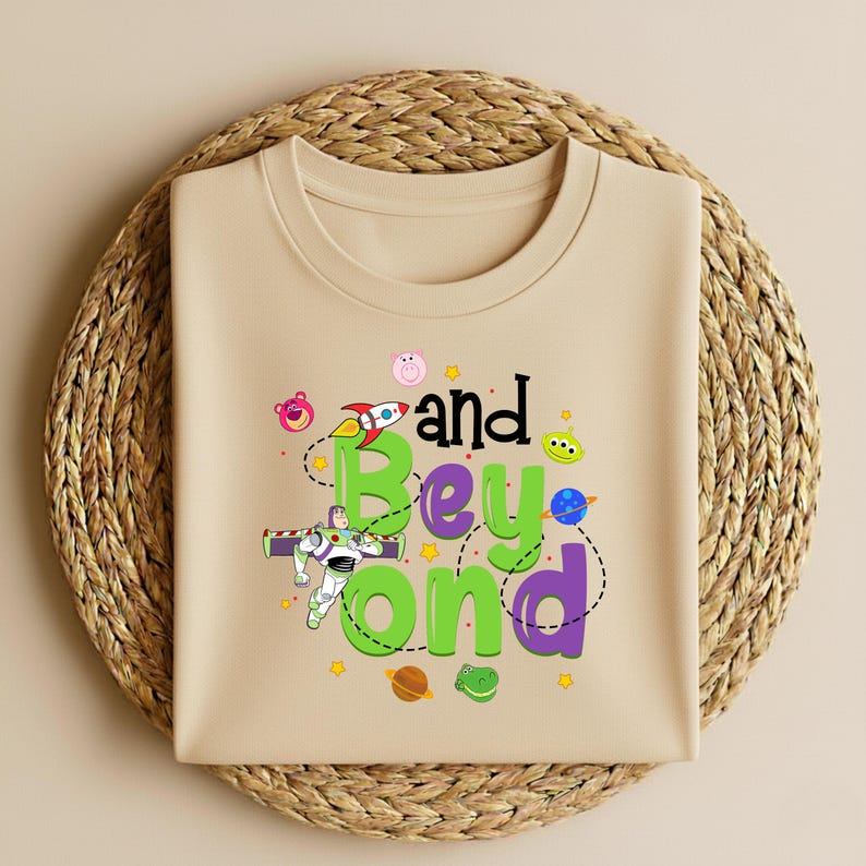 Może przedstawiać: Beżowa koszulka z kolorową grafiką z napisem "and Beyond" i postaciami z kresk&oacute;wek. Projekt zawiera rakietę, zielonego kosmitę i inne elementy o tematyce kosmicznej. Koszulka jest prezentowana na tkanej, okrągłej macie.
