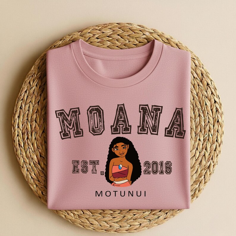 May include: A pale pink sweatshirt featuring the word "MOANA" in a distressed brown font. Below the word is a cartoon illustration of a woman and the text "EST. 2016 MOTUNUI". The sweatshirt is folded on a woven, circular placemat.