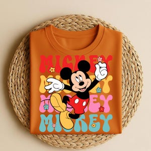 May include: Orange t-shirt featuring a retro design with the text "Mickey" in colorful stacked letters. The graphic includes a cartoon Mickey Mouse character and flower accents. The shirt is folded on a woven mat.