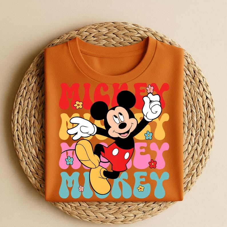 May include: Orange t-shirt featuring a retro design with the text "Mickey" in colorful stacked letters. The graphic includes a cartoon Mickey Mouse character and flower accents. The shirt is folded on a woven mat.