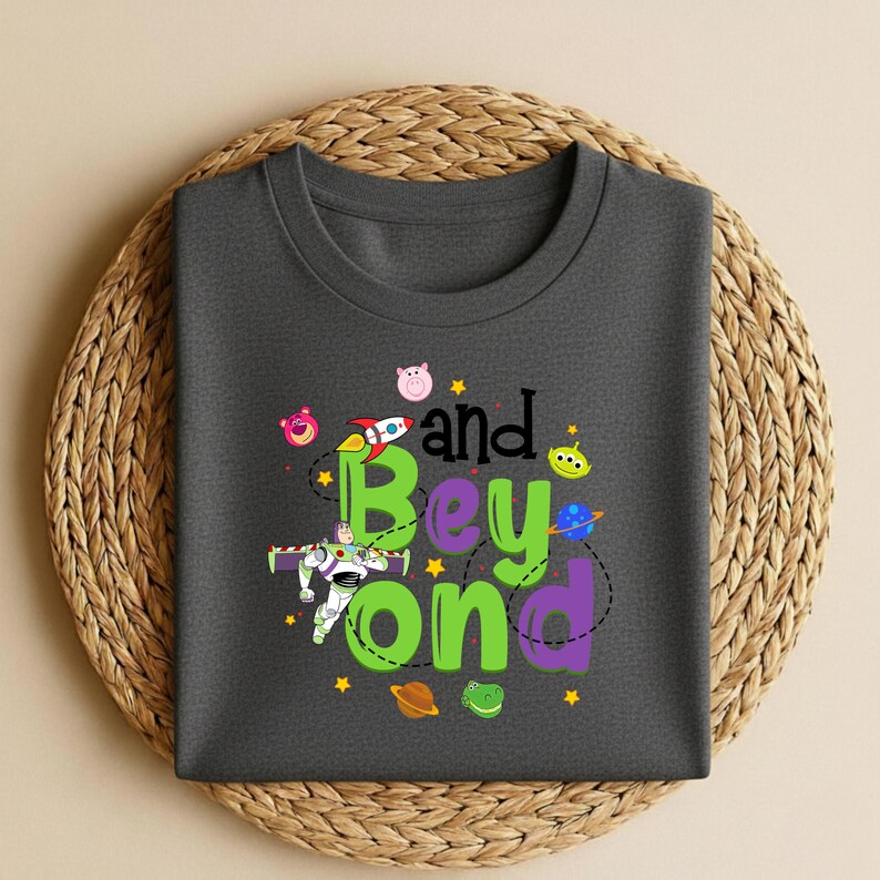 Może przedstawiać: Ciemnoszara koszulka z okrągłym dekoltem z napisem "and Beyond" w kolorze zielonym i fioletowym, z ilustracjami z kresk&oacute;wek przedstawiającymi rakietę, Buzz Lightyear i inne postacie. Koszulka leży na okrągłej, tkanej podkładce.