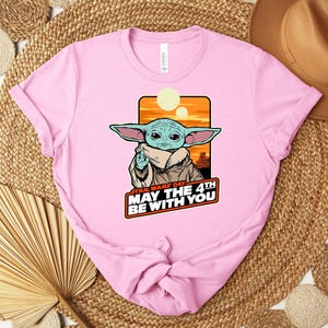 Puede incluir: Camiseta rosa claro con un dise&ntilde;o de Grogu de Star Wars. El dise&ntilde;o incluye el texto "STAR WARS DAY MAY THE 4TH BE WITH YOU" en rojo y blanco. La camiseta est&aacute; sobre una superficie tejida.