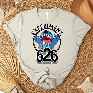 May include: A light beige t-shirt with a graphic of the character Stitch. The design includes the text "EXPERIMENT" in a circular frame, the number "626", and a blue Stitch with an open mouth.