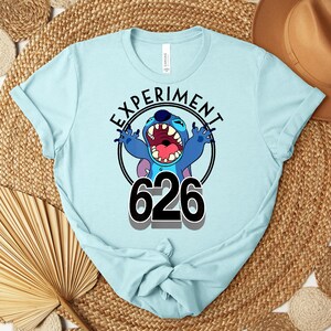 May include: Pale blue t-shirt with a cartoon character and the text "EXPERIMENT 626" in a black circle. The character is blue with large ears and an open mouth. The shirt is made of a soft material.