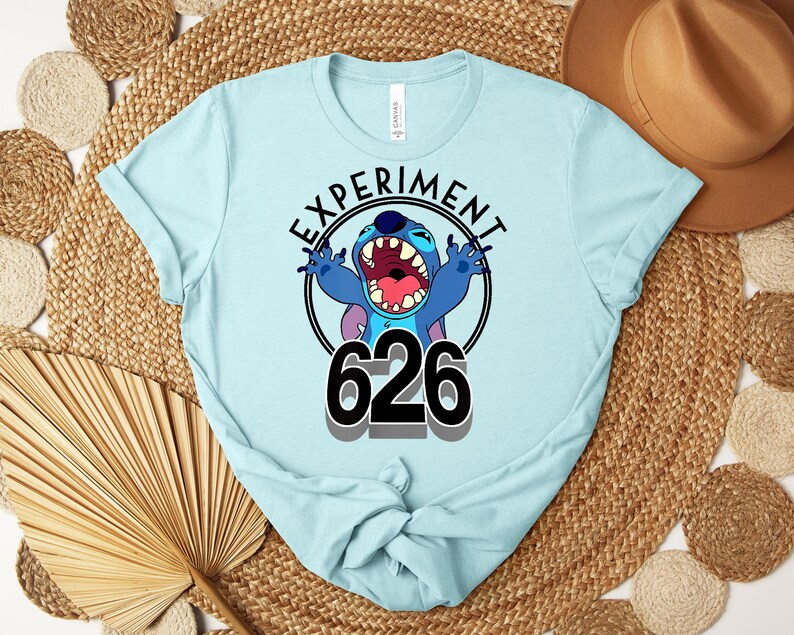 May include: Pale blue t-shirt with a cartoon character and the text "EXPERIMENT 626" in a black circle. The character is blue with large ears and an open mouth. The shirt is made of a soft material.