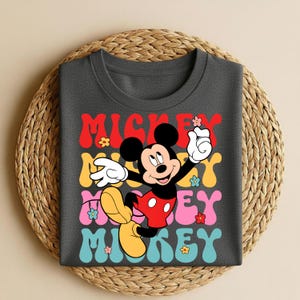 May include: Dark gray t-shirt featuring a colorful graphic of Mickey Mouse with the text "Mickey" in red, yellow, pink, and blue. The design includes flowers and a retro font, creating a playful and nostalgic vibe.