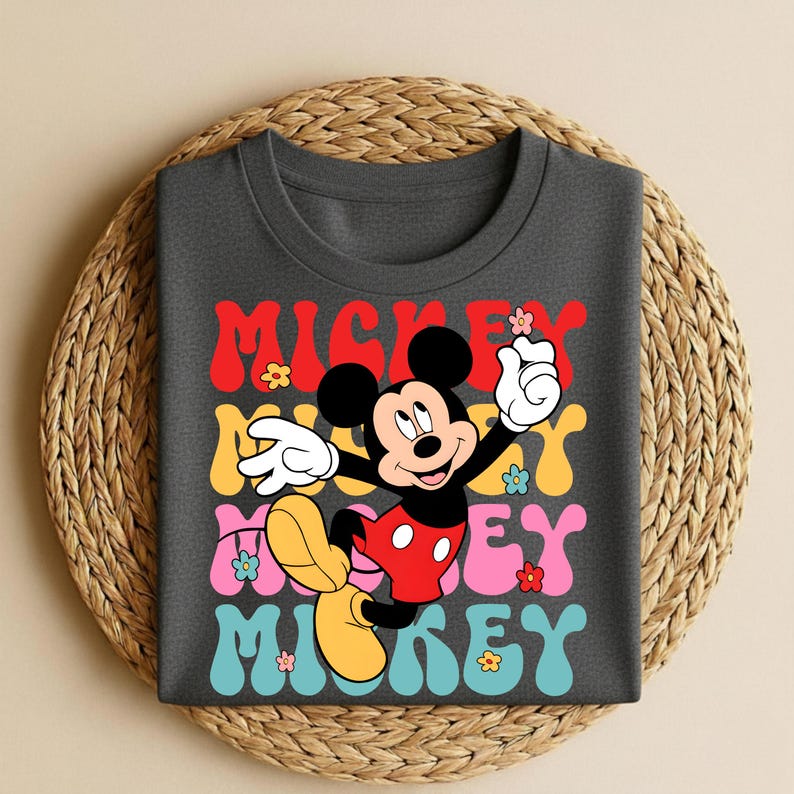 May include: Dark gray t-shirt featuring a colorful graphic of Mickey Mouse with the text "Mickey" in red, yellow, pink, and blue. The design includes flowers and a retro font, creating a playful and nostalgic vibe.