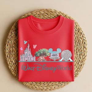 May include: A red crew neck t-shirt featuring a graphic of a castle, tree, film reel, and a sphere, with the text "Walt Disney World" in teal. The design includes Mickey Mouse ears and the word "Studios".