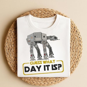 May include: White t-shirt featuring a gray AT-AT walker illustration with the text "GUESS WHAT DAY IT IS?" in a yellow and black rectangular box. The shirt is displayed on a woven, circular placemat.