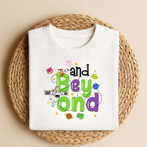 Może przedstawiać: Biała koszulka z napisem "and Beyond" w kolorze zielonym i fioletowym, z kresk&oacute;wkową grafiką rakiety, planet i kosmicznego strażnika. Projekt jest wyśrodkowany na koszulce.