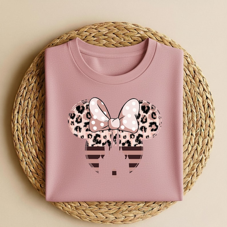 May include: A dusty rose-colored sweatshirt featuring a Minnie Mouse design. The design includes a bow with white polka dots, leopard print ears, and a striped face. The sweatshirt is folded on a woven, circular placemat.