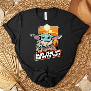 Puede incluir: Camiseta negra con un gr&aacute;fico de un personaje alien&iacute;gena verde con orejas grandes, sobre un fondo naranja y amarillo. El texto "STAR WARS DAY MAY THE 4TH BE WITH YOU" se muestra debajo en rojo y blanco.