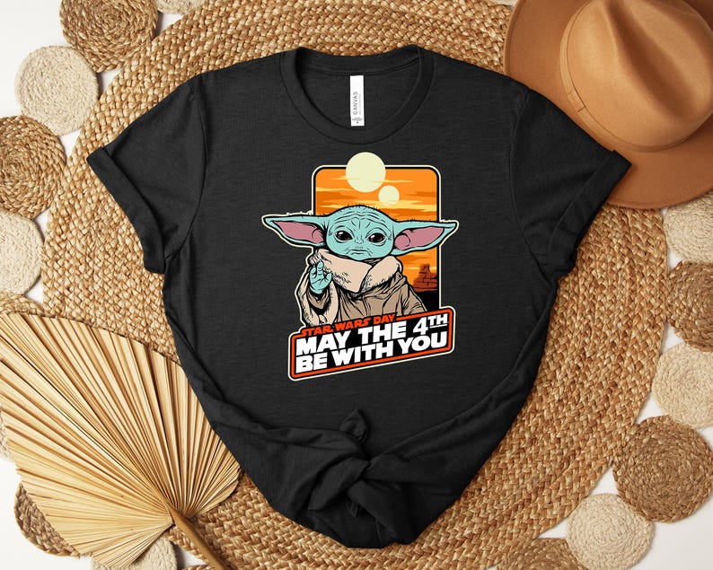 Puede incluir: Camiseta negra con un gr&aacute;fico de un personaje alien&iacute;gena verde con orejas grandes, sobre un fondo naranja y amarillo. El texto "STAR WARS DAY MAY THE 4TH BE WITH YOU" se muestra debajo en rojo y blanco.