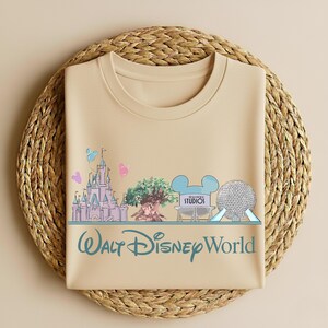 May include: Beige t-shirt featuring a Walt Disney World design. The design includes a castle, tree, Mickey Mouse ears, and a silver sphere. The text "Walt Disney World" is printed in teal.
