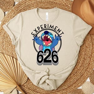 May include: Beige t-shirt with a cartoon character in a black circle, with the text "EXPERIMENT" above and the number "626" below. The character is blue with large ears and an open mouth. The shirt has a crew neck and short sleeves.