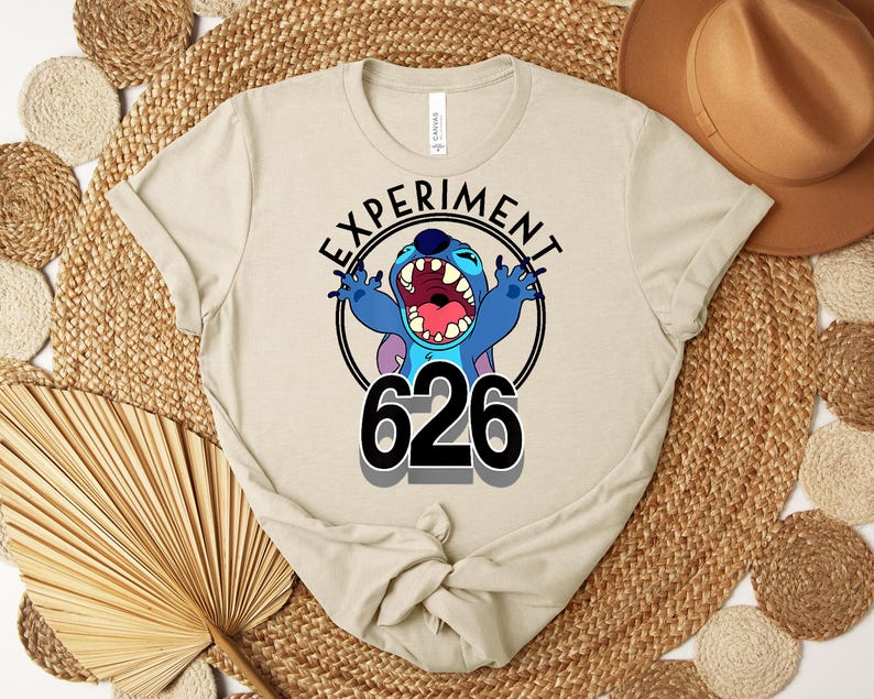 May include: Beige t-shirt with a cartoon character in a black circle, with the text "EXPERIMENT" above and the number "626" below. The character is blue with large ears and an open mouth. The shirt has a crew neck and short sleeves.