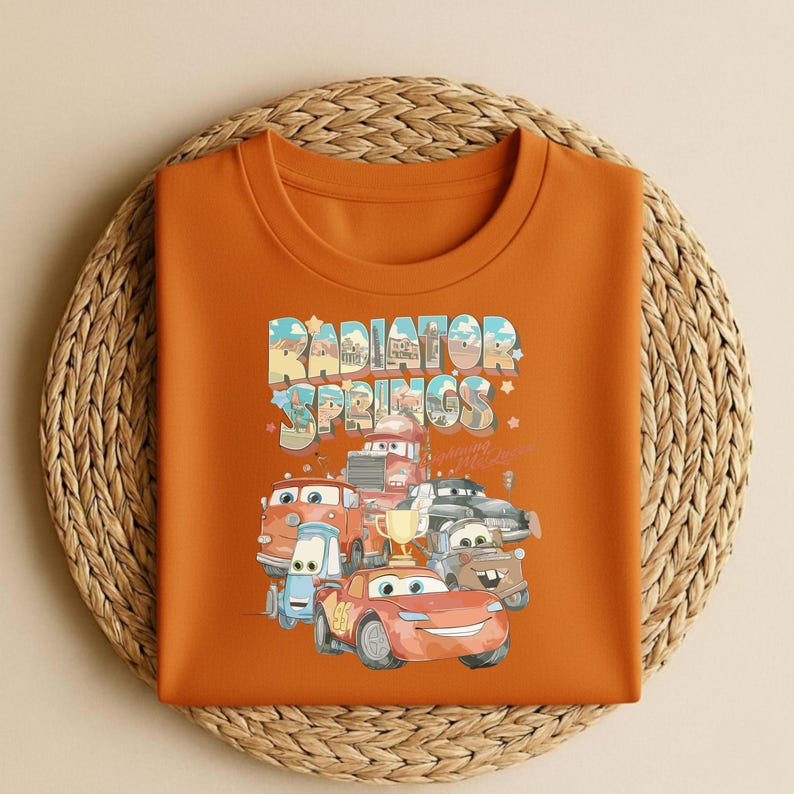 May include: Orange crew neck sweatshirt featuring a colorful graphic of characters from the movie Cars, with the text "Radiator Springs" in a playful font. The design includes various car characters and a trophy.