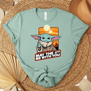 Puede incluir: Camiseta verde claro con un gr&aacute;fico de un personaje alien&iacute;gena verde con orejas grandes, sobre un fondo naranja y amarillo. El texto "STAR WARS DAY MAY THE 4TH BE WITH YOU" se muestra debajo del personaje. La camiseta est&aacute; anudada en la parte inferior.