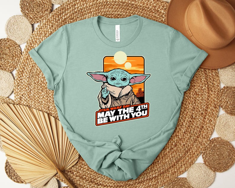 Puede incluir: Camiseta verde claro con un gr&aacute;fico de un personaje alien&iacute;gena verde con orejas grandes, sobre un fondo naranja y amarillo. El texto "STAR WARS DAY MAY THE 4TH BE WITH YOU" se muestra debajo del personaje. La camiseta est&aacute; anudada en la parte inferior.