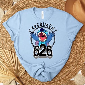 May include: Pale blue t-shirt with a cartoon character and the text "EXPERIMENT 626" in a black circle. The character is blue with large ears and an open mouth. The shirt is made of a soft material.