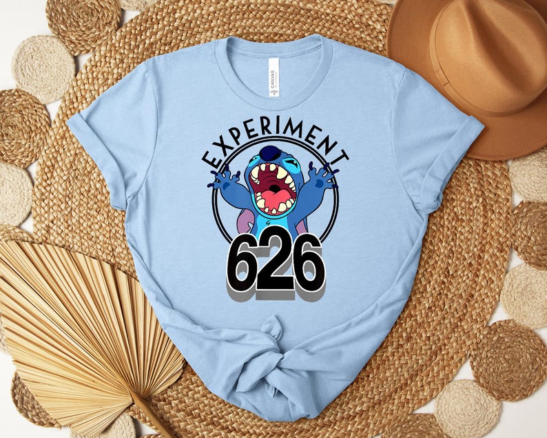 May include: Pale blue t-shirt with a cartoon character and the text "EXPERIMENT 626" in a black circle. The character is blue with large ears and an open mouth. The shirt is made of a soft material.