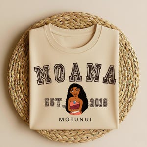 May include: A beige t-shirt featuring the word "MOANA" in a brown, collegiate-style font. Below the word "MOANA" is a cartoon illustration of a woman, the text "EST. 2016" and the word "MOTUNUI". The t-shirt is on a woven, circular placemat.
