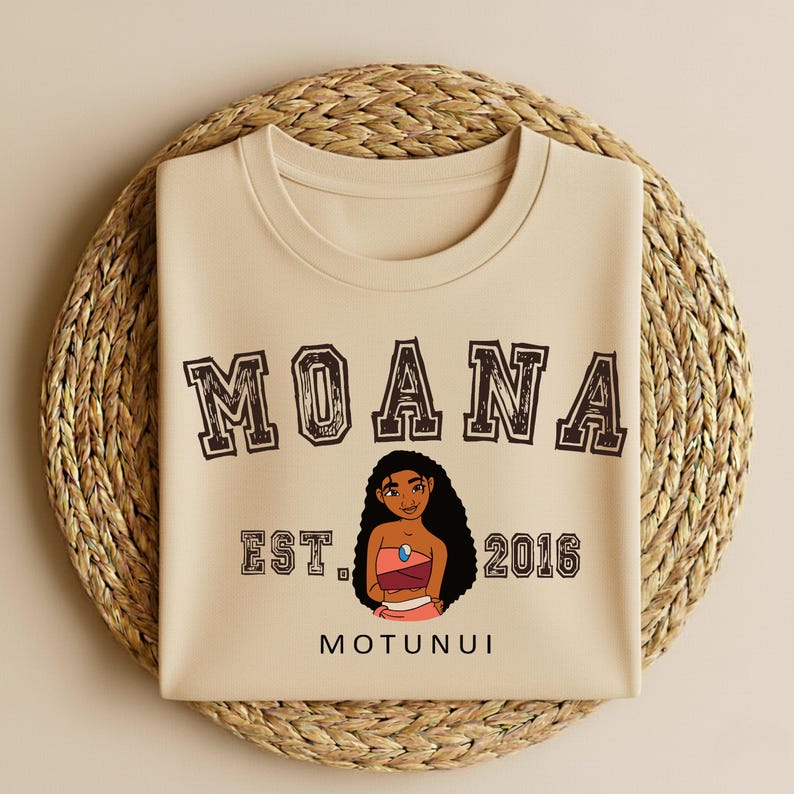 May include: A beige t-shirt featuring the word "MOANA" in a brown, collegiate-style font. Below the word "MOANA" is a cartoon illustration of a woman, the text "EST. 2016" and the word "MOTUNUI". The t-shirt is on a woven, circular placemat.