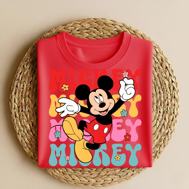 May include: A red t-shirt featuring a colorful graphic of Mickey Mouse with the word "Mickey" in a retro font. The design includes flowers and a playful pose, perfect for Disney fans.
