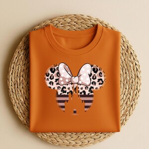 May include: An orange t-shirt featuring a Minnie Mouse design. The design includes a bow with white polka dots, leopard print ears, and a castle silhouette. The shirt is folded on a woven mat.