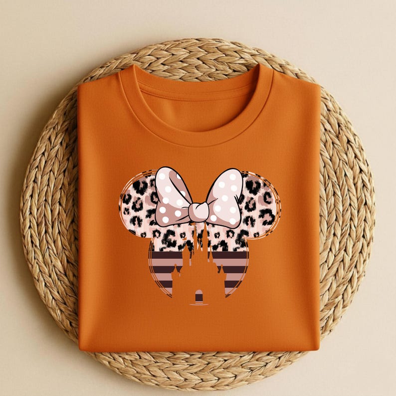 May include: An orange t-shirt featuring a Minnie Mouse design. The design includes a bow with white polka dots, leopard print ears, and a castle silhouette. The shirt is folded on a woven mat.