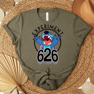 May include: Olive green t-shirt with a cartoon character design. The text "EXPERIMENT" is above the character, and "626" is below. The character is blue with large ears and an open mouth. The shirt is made of a soft material.