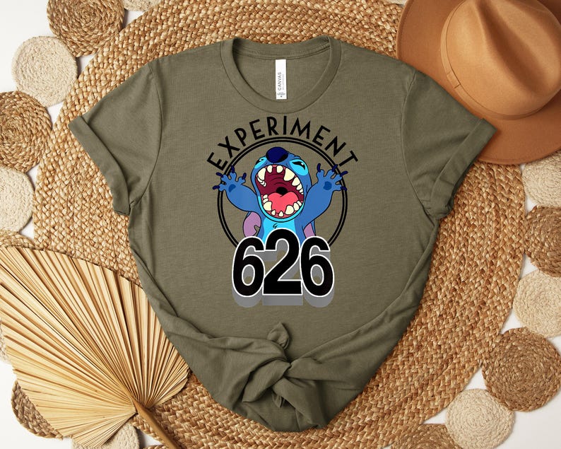 May include: Olive green t-shirt with a cartoon character design. The text "EXPERIMENT" is above the character, and "626" is below. The character is blue with large ears and an open mouth. The shirt is made of a soft material.