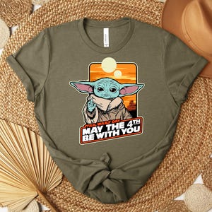Puede incluir: Camiseta verde oliva con un gr&aacute;fico de un personaje con orejas grandes y el texto "STAR WARS DAY MAY THE 4TH BE WITH YOU". El gr&aacute;fico incluye una puesta de sol naranja y dos lunas.