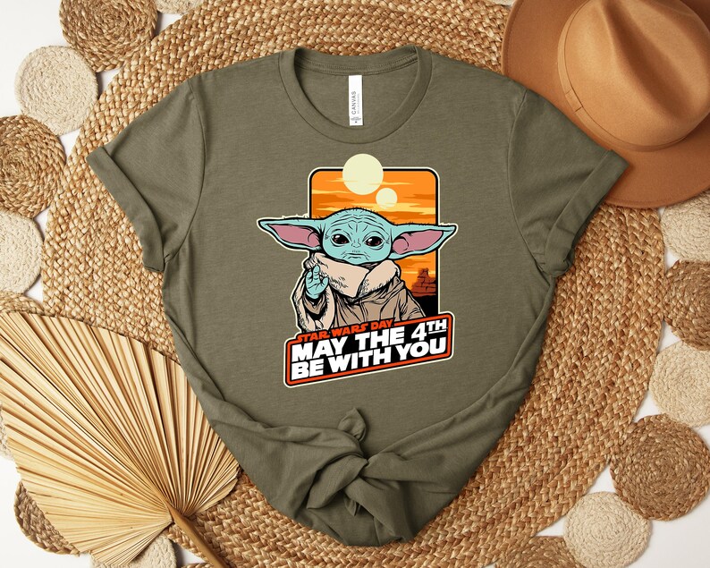 Puede incluir: Camiseta verde oliva con un gr&aacute;fico de un personaje con orejas grandes y el texto "STAR WARS DAY MAY THE 4TH BE WITH YOU". El gr&aacute;fico incluye una puesta de sol naranja y dos lunas.