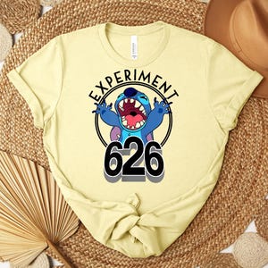 May include: A pale yellow t-shirt with a graphic of the character Stitch from Lilo & Stitch. The design includes the text "EXPERIMENT" above the character and the number "626" below. The character is blue with an open mouth.
