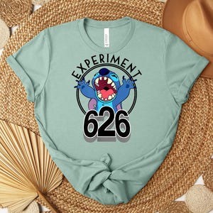May include: A pale green t-shirt with a graphic of the character Stitch. The design includes the text "EXPERIMENT" above the character and the number "626" below. The character is blue with large teeth and is in a screaming pose.