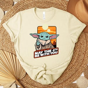 Puede incluir: Camiseta color crema con un gr&aacute;fico de Grogu de Star Wars, con el texto "Star Wars Day May The 4th Be With You". El dise&ntilde;o incluye un fondo de puesta de sol naranja y amarillo.