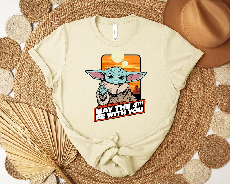 Puede incluir: Camiseta color crema con un gr&aacute;fico de Grogu de Star Wars, con el texto "Star Wars Day May The 4th Be With You". El dise&ntilde;o incluye un fondo de puesta de sol naranja y amarillo.