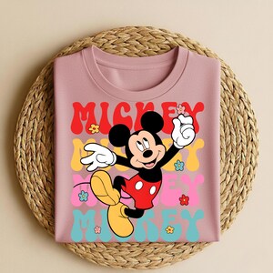 May include: A pink t-shirt featuring a graphic of Mickey Mouse with the text "Mickey" in red, yellow, pink, and blue. The design includes flowers and a retro font. The shirt is folded on a woven mat.