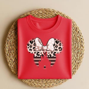 May include: A red crew neck sweatshirt featuring a Minnie Mouse design. The design includes a bow with white polka dots, leopard print ears, and a striped castle silhouette. The sweatshirt is folded on a woven mat.