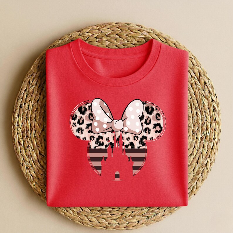 May include: A red crew neck sweatshirt featuring a Minnie Mouse design. The design includes a bow with white polka dots, leopard print ears, and a striped castle silhouette. The sweatshirt is folded on a woven mat.