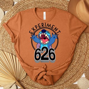 May include: A rust-coloured t-shirt featuring a cartoon image of Stitch with the text "EXPERIMENT" above and "626" below. Stitch is blue with an open mouth and raised arms. The shirt is folded at the bottom.