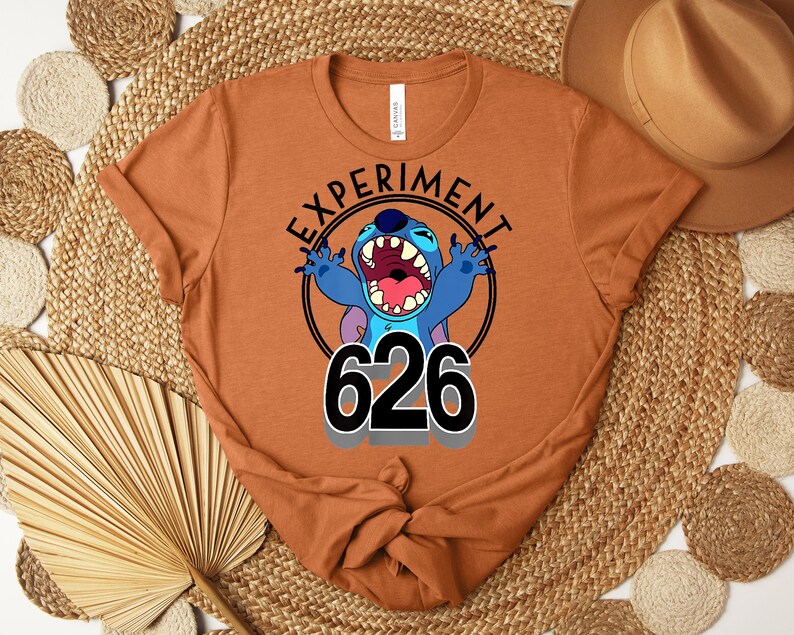 May include: A rust-coloured t-shirt featuring a cartoon image of Stitch with the text "EXPERIMENT" above and "626" below. Stitch is blue with an open mouth and raised arms. The shirt is folded at the bottom.
