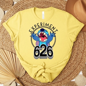May include: A yellow t-shirt with a graphic of Stitch from Lilo & Stitch. The design includes the text "EXPERIMENT" in a circle around Stitch, with the number "626" below. The shirt is made of a soft material.