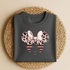 May include: Dark gray t-shirt featuring a Minnie Mouse design. The design includes a bow with pink polka dots, leopard print ears, and a striped castle silhouette. The shirt is displayed on a woven, circular placemat.