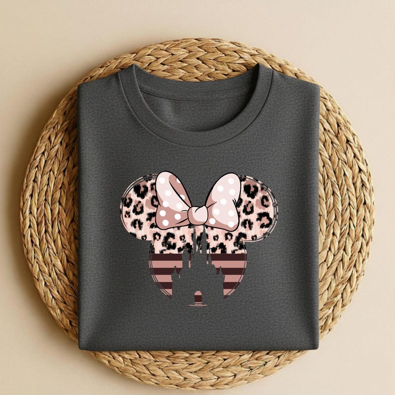 May include: Dark gray t-shirt featuring a Minnie Mouse design. The design includes a bow with pink polka dots, leopard print ears, and a striped castle silhouette. The shirt is displayed on a woven, circular placemat.