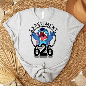 May include: Light grey t-shirt with the text "EXPERIMENT 626" and a cartoon character. The character is blue with large ears and an open mouth. The text is in a black circle.