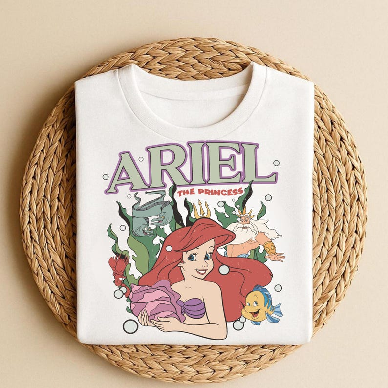 Puede incluir: Sudadera blanca de cuello redondo con un gr&aacute;fico colorido de Ariel de La Sirenita. El gr&aacute;fico incluye la palabra "ARIEL" en verde y morado, junto con otros personajes de la pel&iacute;cula. La sudadera se muestra sobre una estera tejida.