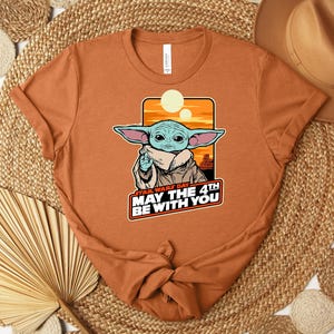 Puede incluir: Camiseta de color &oacute;xido con un gr&aacute;fico de Grogu de Star Wars. La imagen incluye el texto "Star Wars Day May the 4th Be With You". El dise&ntilde;o muestra a Grogu con orejas grandes y una capa beige sobre un fondo de puesta de sol naranja.