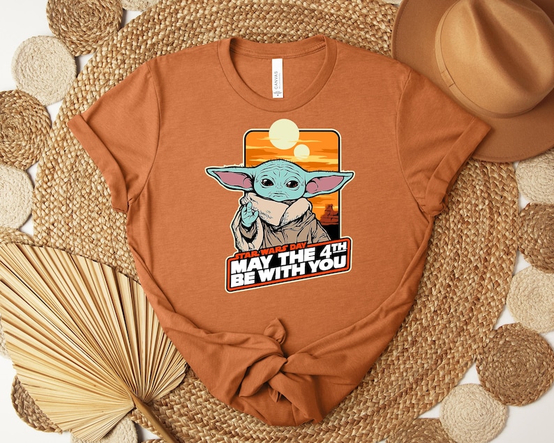 Puede incluir: Camiseta de color &oacute;xido con un gr&aacute;fico de Grogu de Star Wars. La imagen incluye el texto "Star Wars Day May the 4th Be With You". El dise&ntilde;o muestra a Grogu con orejas grandes y una capa beige sobre un fondo de puesta de sol naranja.